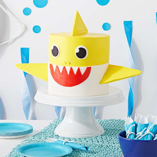 Baby Shark cake
Start by stacking four cakes with frosting between each layer. For the mouth – roll out white fondant and place onto the cake. Then, roll out a red Fruit Roll-Up, cut it into the shape of a mouth and place it onto the white fondant. For the eyes – cut out two circles from the white fondant, then, cut out two smaller circles from black fondant and place them onto the white eyes. For the fins – bake triangle sugar cookies, and using wooden dowels, insert into the sides and top of the cake.

Tip: Place the dowels/skewers in the fin cookies before they bake.
