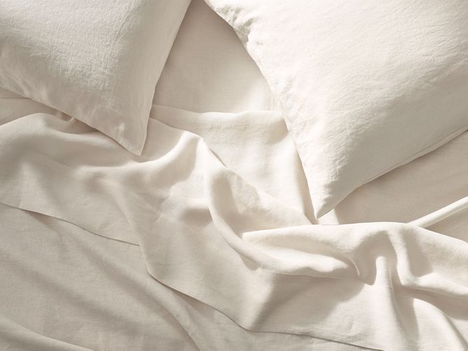Casaluna bedding & bath. Natural colored, 100% linen pillowcases and sheets viewed from above on a softly lit, loosely made bed. Right image features a stack of natural and light gray towels.