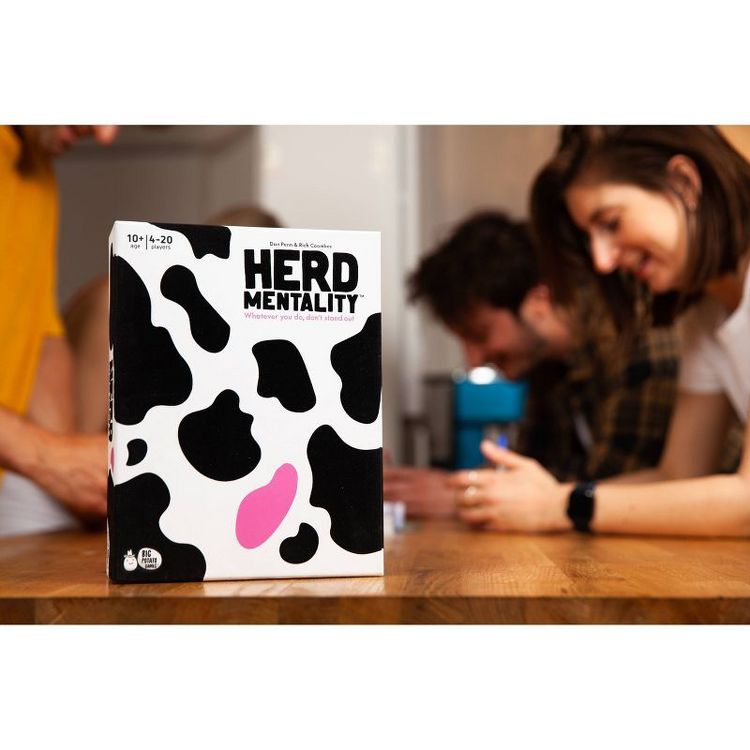 Box of "Herd Mentality" game with a black and white cow pattern and a pink spot on a wooden table.