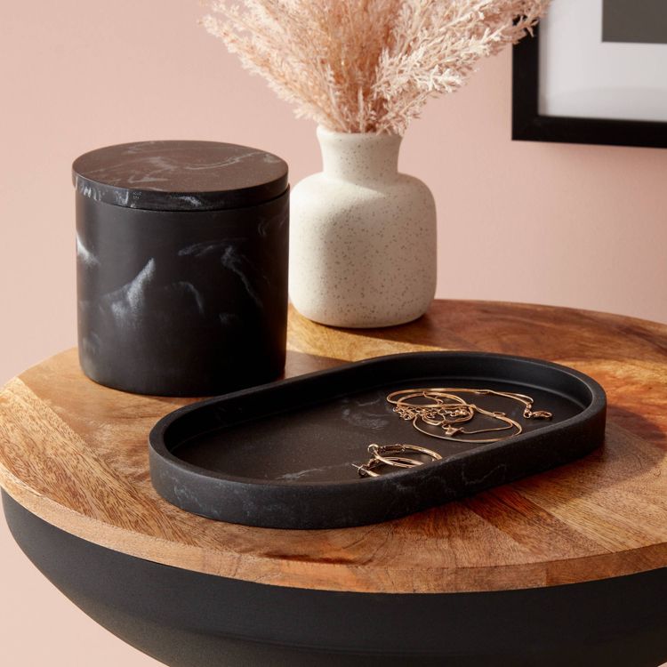 A black colored oval tray with jewelry placed on a wooden surface next to a black container and a white vase.