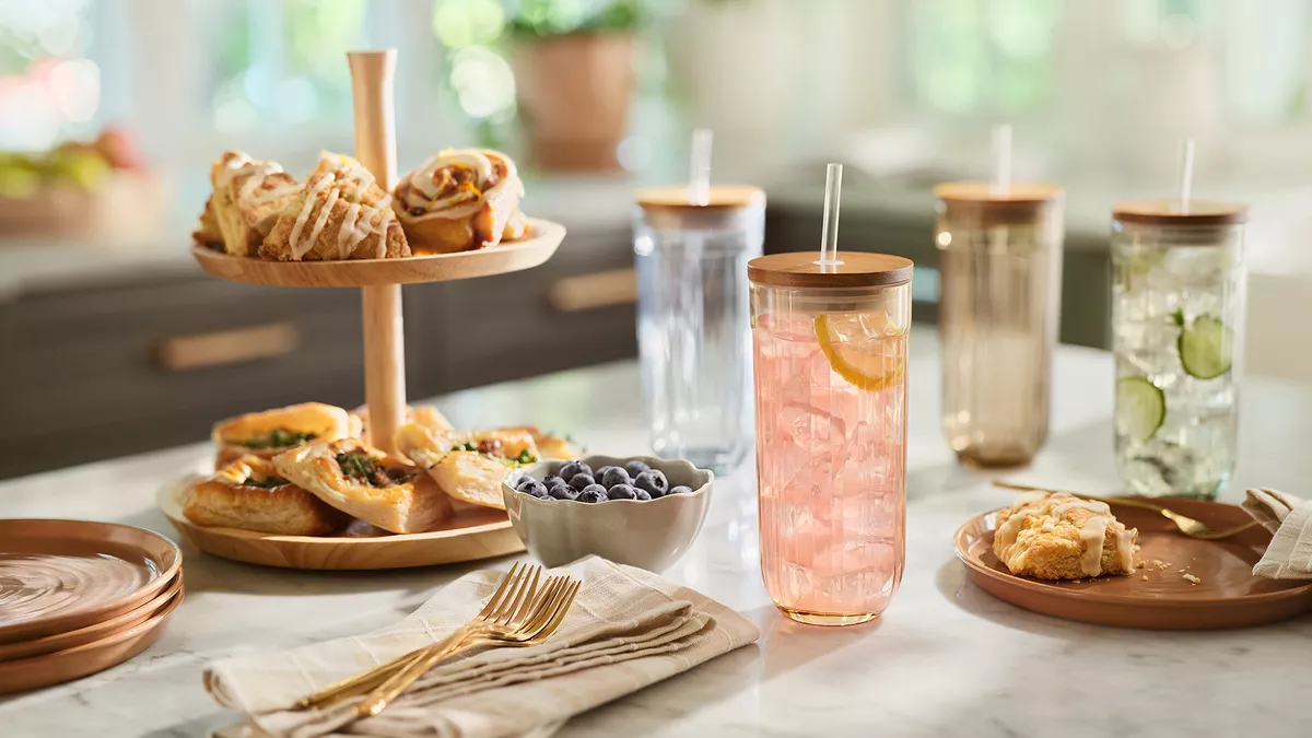 Glass tumblers filled with icy drinks on a kitchen countertop, and a pitcher of a pink liquid atop a coffee table  set with light snacks.