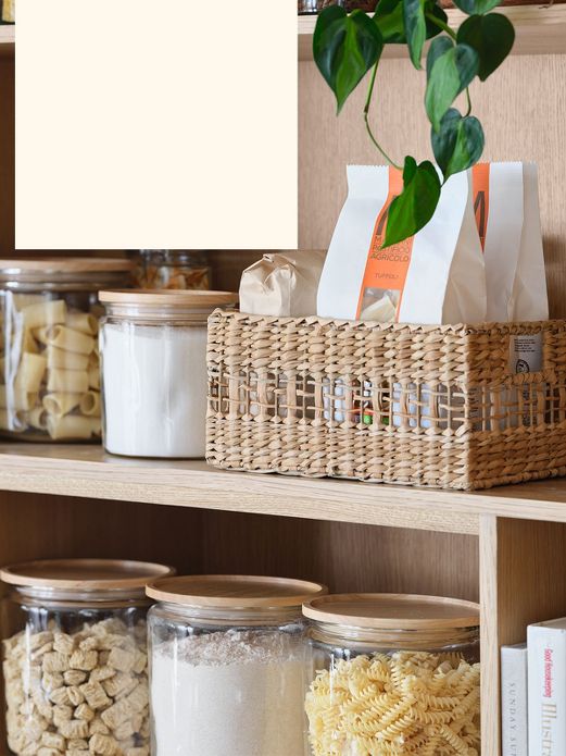 Wicker basket and various glass jars containing dried pasta and baking essentials in pantry.