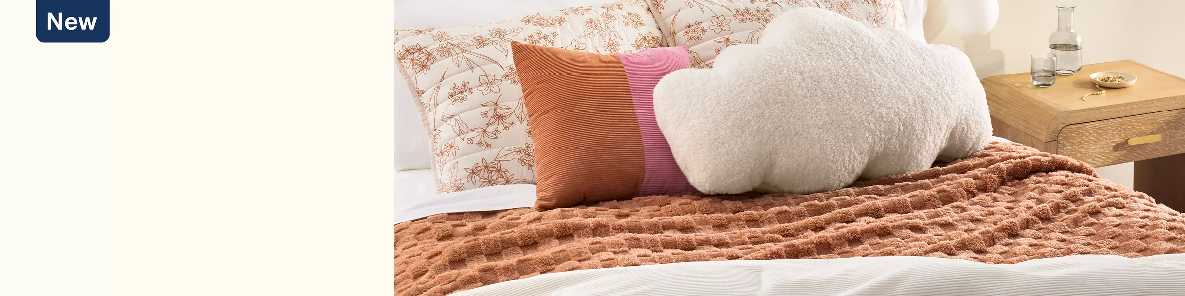 An orange checkered blanket lays on a white comforter. 2 big white pillows with orange florals are at the bed top. An orange & pink color-blocked pillow & a fluffy cloud-shaped pillow sit in front. Warm colors & a mix of textures make a cozy room.  