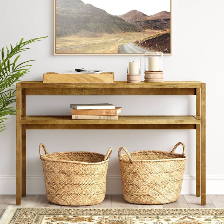 A wooden console table with two shelves, holding books, candles, and a tray, with two woven baskets underneath.