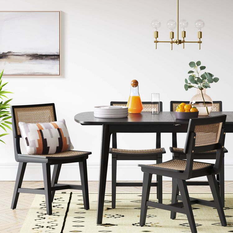 Black colored dining chairs with woven backs and seats are arranged around a dark table, with a decorative pillow on one chair and a light-colored rug underneath.