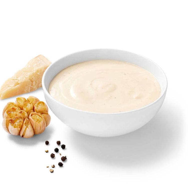 A white bowl filled with creamy light beige colored sauce, accompanied by a piece of cheese, roasted garlic, and black peppercorns.
