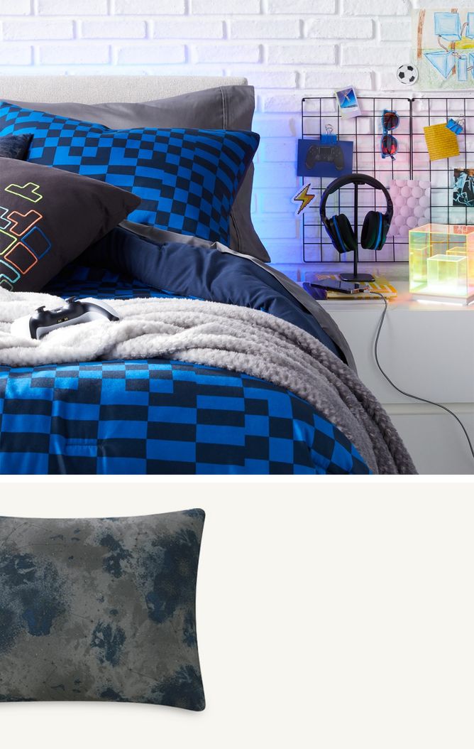 A teen’s bedroom with a white brick accent wall & contrasting dark-colored bedding has layered graphic pillows & a gaming controller on the bed. The side table has a charging cell phone, funky acrylic LED light, headphones & an open bag of chips.