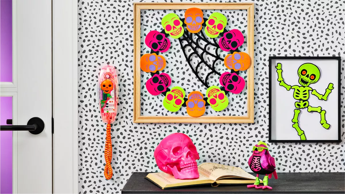 This decor is bright, fun & reflective of the 80s. A pumpkin, pink skull & small purple bird with neon green accessories sit on a side table. A wreath of multi-colored skulls hangs on the wall between a framed skeleton & a Halloween wall phone.