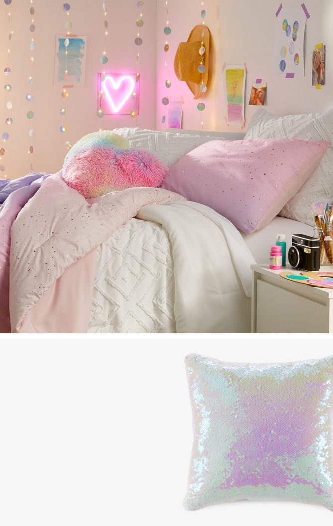 A pastel fur heart pillow lays on a cozy bed of a teen room full of pastel colors & shimmering details. DIY artwork & photos are taped to the walls & a heart-shaped neon sign is lit. A used paint palette, brushes & camera sit on the side table.