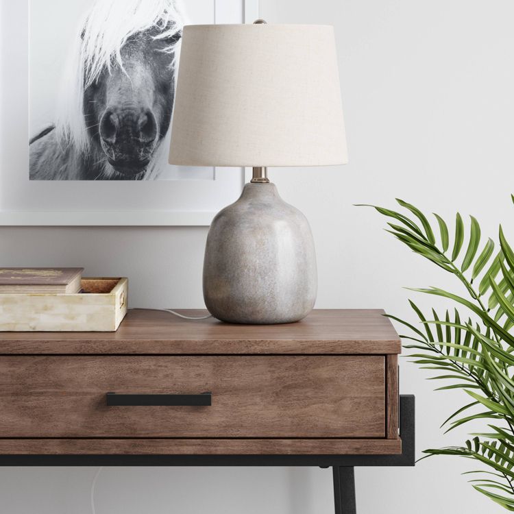 A gray colored table lamp with a beige colored shade sits on a wooden table next to a framed black and white horse photo and a green plant.