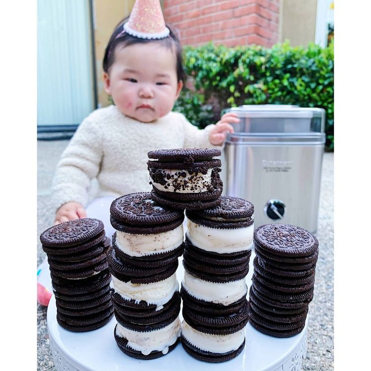 A stainless steel ice cream maker is placed on the ground behind stacks of chocolate sandwich cookies filled with ice cream.