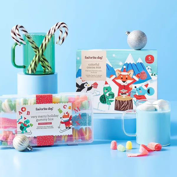 Festive display with holiday treats and drinks on blue stands. A clear box of colorful gummies, a package of colorful cocoa mix with animal illustrations, and a red-striped box of chocolate truffles. Two mugs with creamy drinks and candy canes adorn the scene.