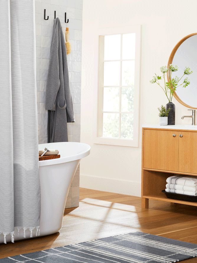 A grey robe hangs on a hook in a bathroom. Accents such as a grey tasseled shower curtain, wooden bath tray and various vases fill the space.