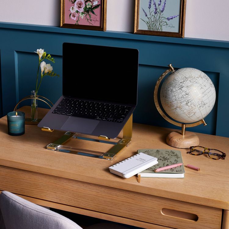 A laptop is placed on a clear and gold colored riser on a wooden desk, surrounded by a globe, notebooks, a pen, glasses, a candle, and a vase with flowers.