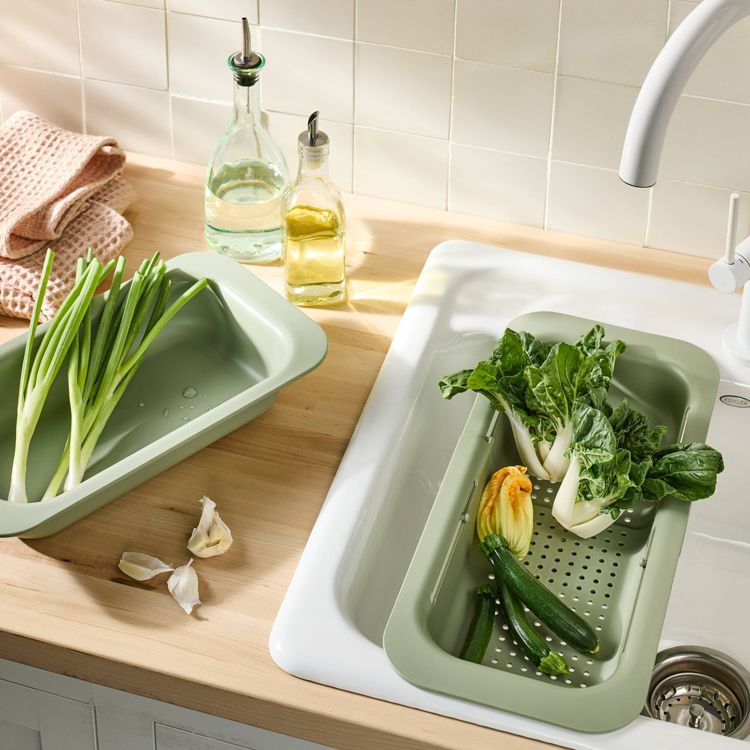 A green colored colander placed over a sink, holding fresh vegetables including zucchini and leafy greens.