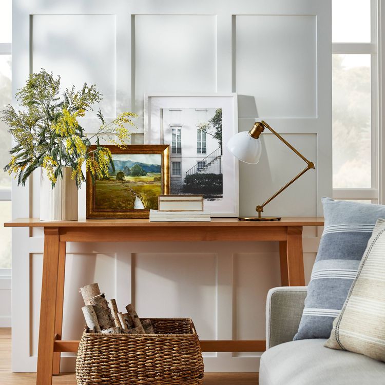 A wooden table with framed artwork, a white lamp, a vase with yellow flowers, and a basket of logs underneath, set against a paneled wall.