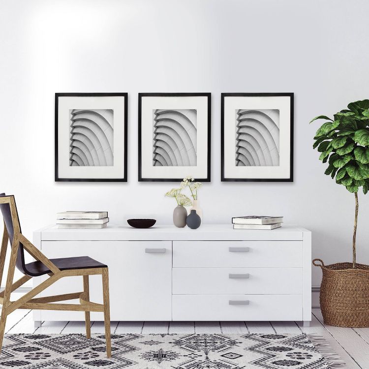 Three black-framed abstract art pieces hang above a white sideboard with books, vases, and a plant, next to a wooden chair on a patterned rug.