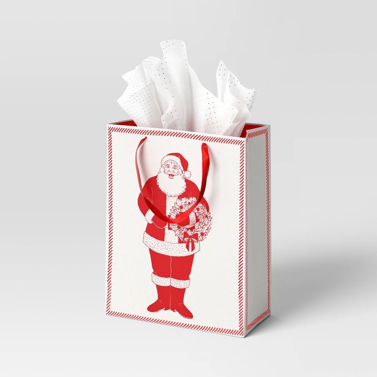 A white colored gift bag with a red Santa Claus design and red handles, filled with white tissue paper.