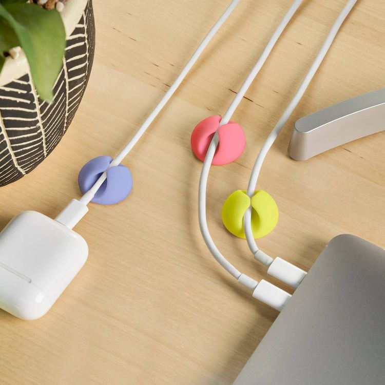 Three colored cable clips in purple, pink, and yellow hold white charging cables on a wooden surface.
