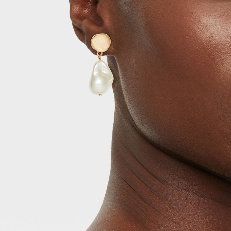 A gold colored earring with a pearl-like drop is worn on an ear.