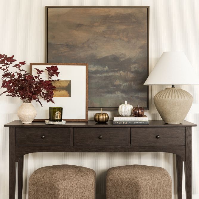 A dark wood console table decorated with moody artwork, a stone vase and lamp, and marble pumpkin decor. Two geometric ottomans are tucked beneath.