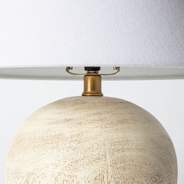 A table lamp with a textured beige colored base and a white colored fabric shade, featuring a brass colored neck.