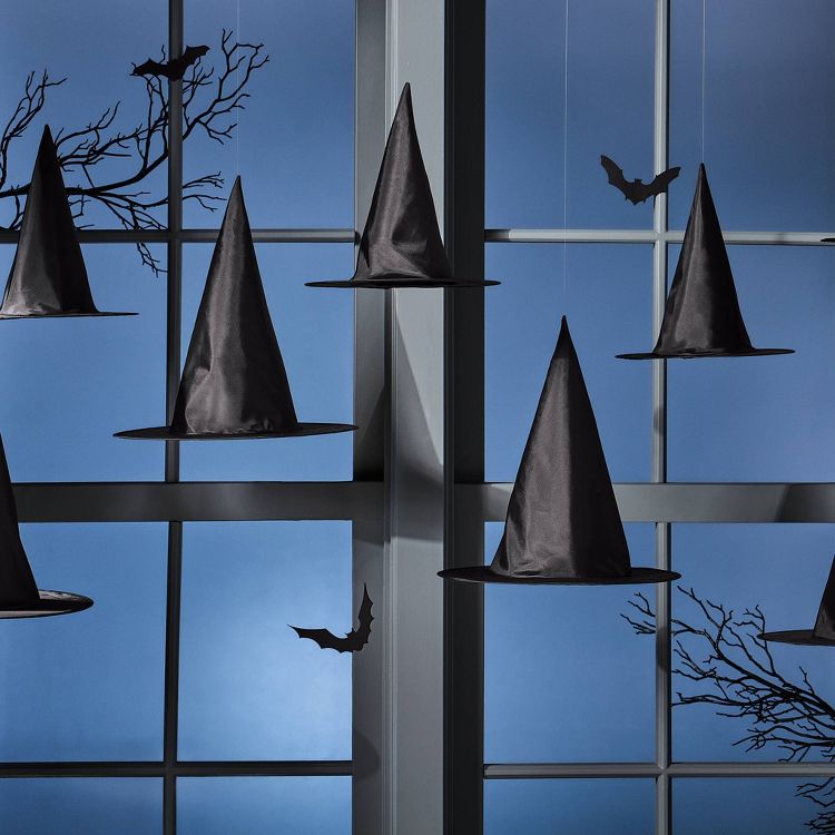 Black colored witch hats and bat silhouettes hanging in front of a window with a blue background.