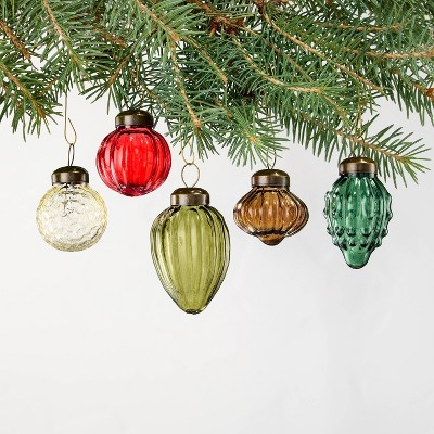 Five glass ornaments in red, yellow, green, brown, and dark green colors hang from a pine branch.