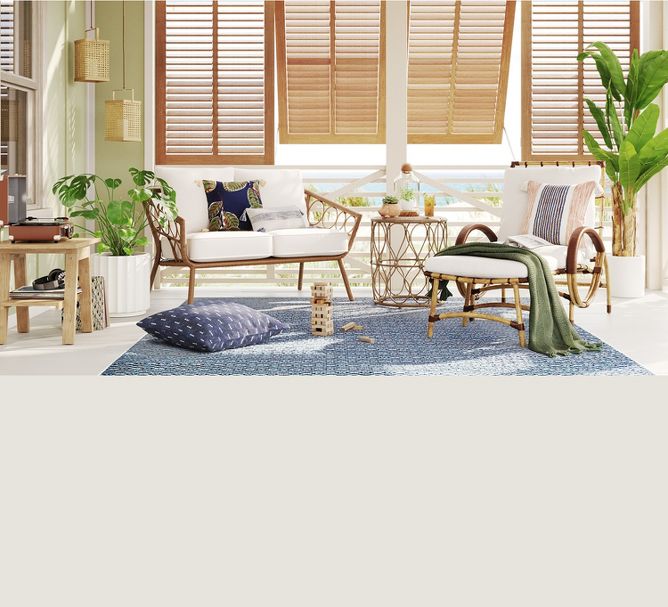A patio with cushioned wicker chairs, printed pillows & a table with succulents sit on a graphic rug. A blanket & book are in one chair & a pillow & Jenga game lay on the floor. Tall plants frame the space & open shutters create a bright & airy vibe. 