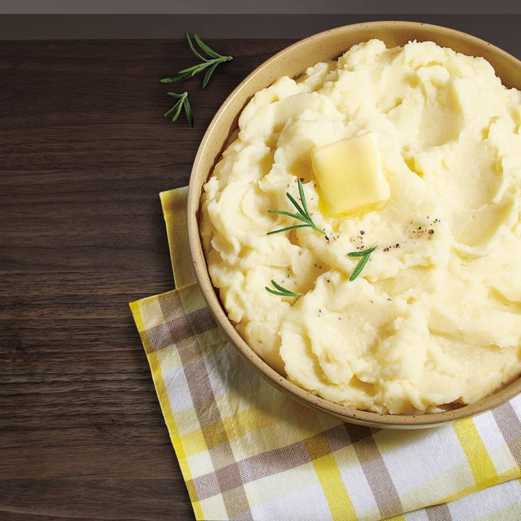 A bowl of mashed potatoes topped with a pat of butter and garnished with rosemary, placed on a yellow and white checkered cloth on a wooden surface.