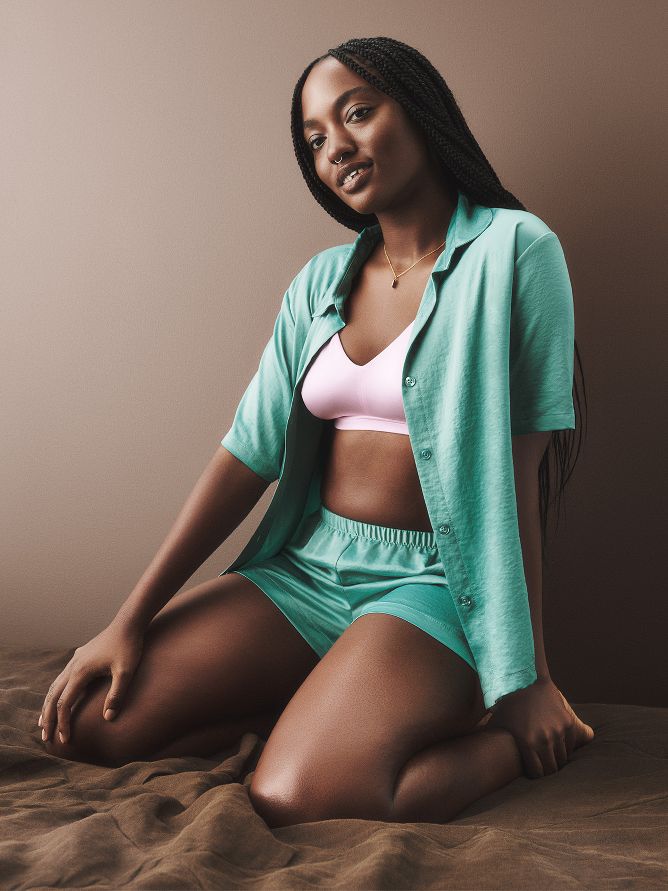 A woman is wearing a silky aqua green pajama set of shorts and a matching button-up short-sleeve shirt. Underneath, she adds a pop of contrasting color with a pink bralette.