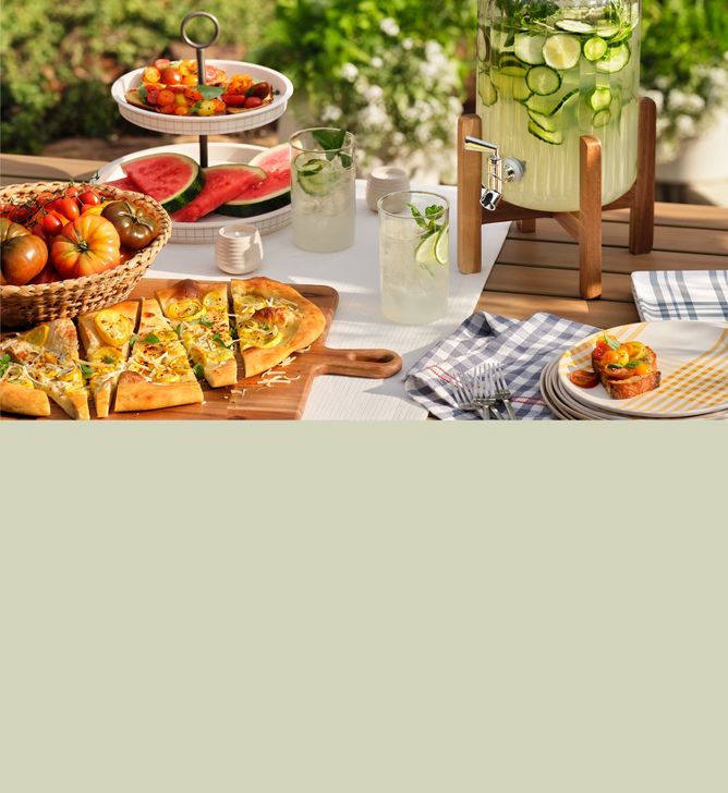 Outdoor snack table features tiered tray, flatbread on wood paddle serve board and lemonade in drink dispenser. 