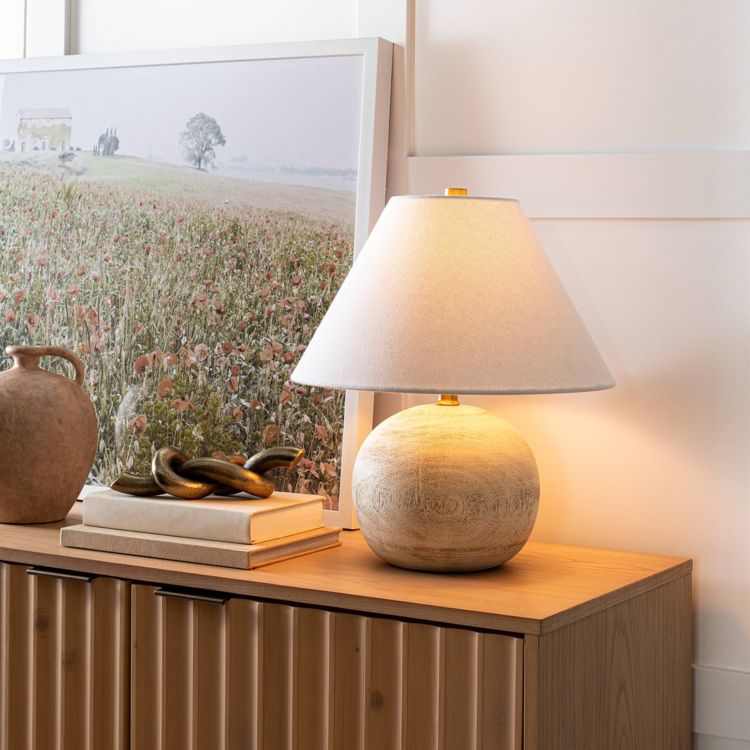 A table lamp with a round, light-colored base and a white colored shade is placed on a wooden cabinet next to a framed landscape picture and decorative items.