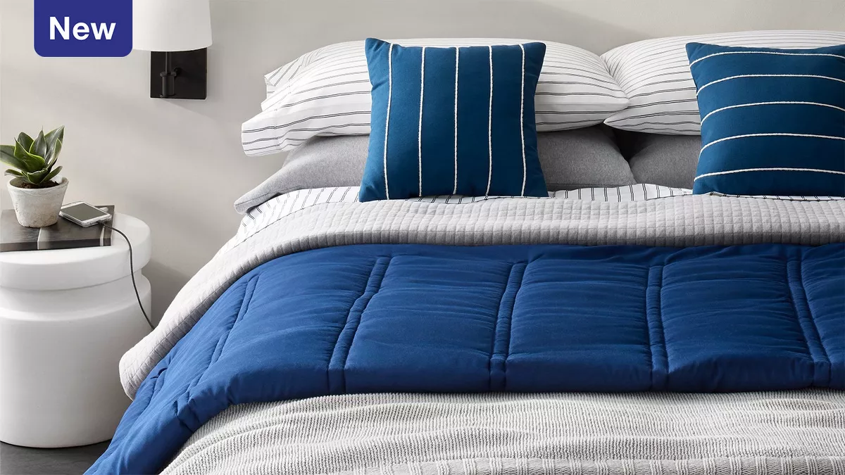 Light gray pillows & white & gray striped pillows are stacked on a bed. 2 navy & white striped accent pillows lean on the stack. A knit gray blanket & navy plush comforter cover the bed. The cool, contrasting tones & textures make a crisp & neat room. 