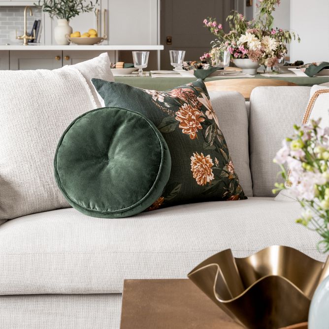 Velvety dark green pillows on a white couch. One is round while the other is square with a pink floral graphic print. A gold toned ruffle bowl is out of focus in the forefront.