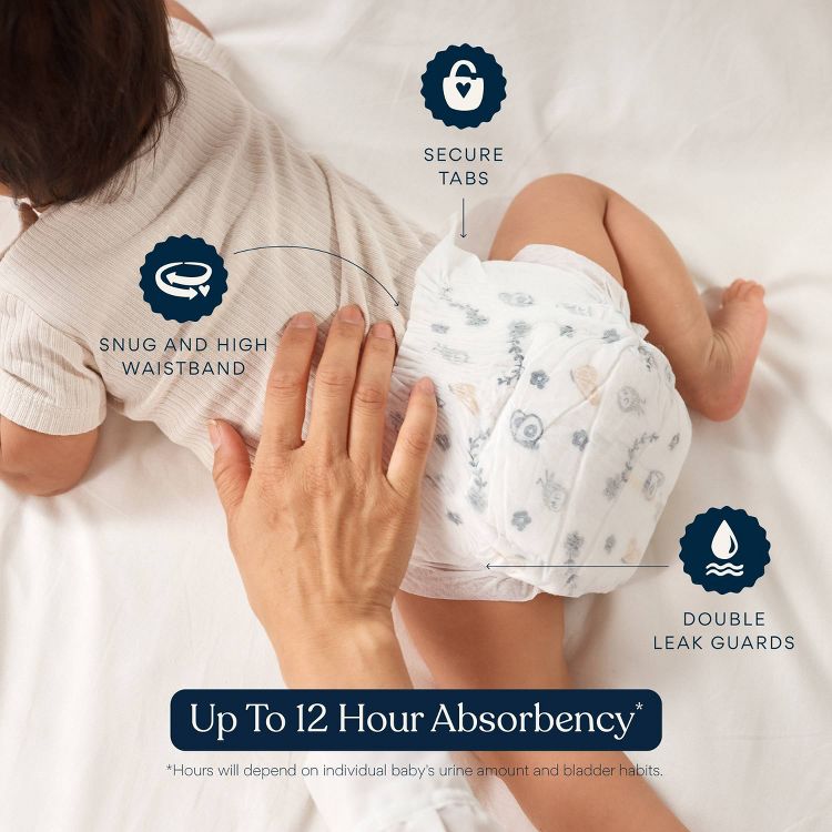 A baby wearing a white colored diaper with blue and beige patterns, featuring secure tabs, a snug and high waistband, and double leak guards, with text stating "Up To 12 Hour Absorbency."
