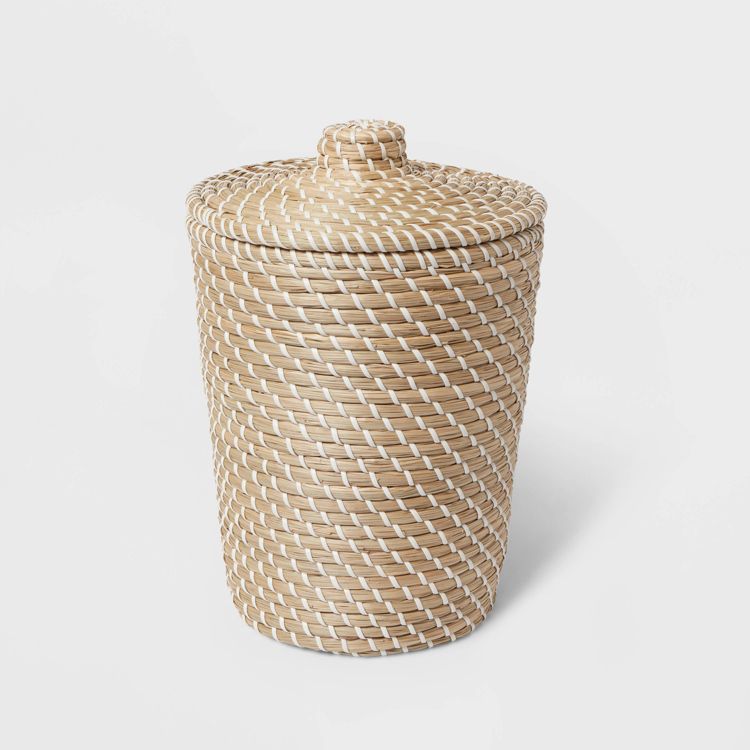 A beige and white colored woven wastebasket with a lid on a white background.