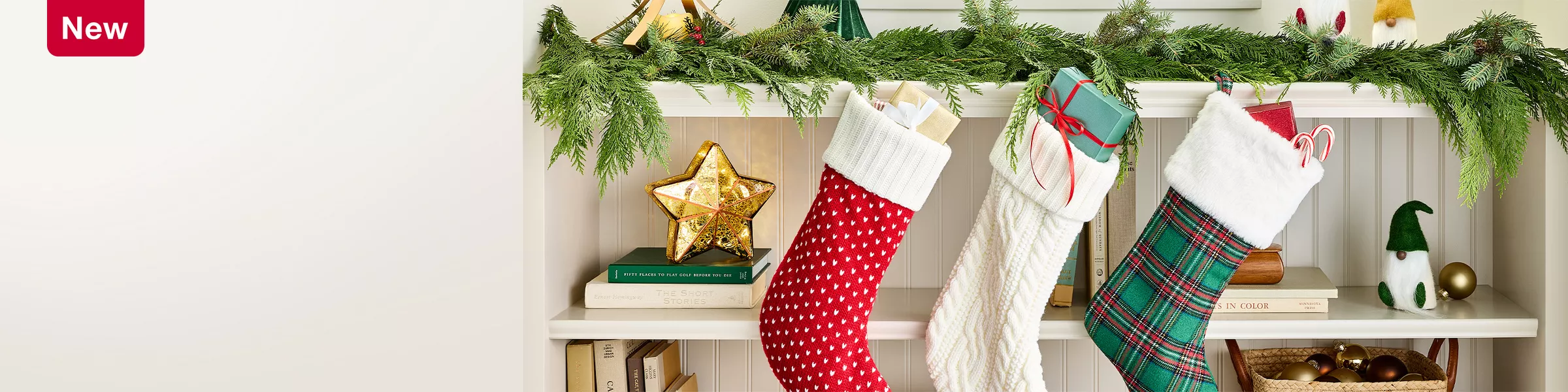 New. A cozy holiday scene with three stockings hung from a shelf, filled with gifts. The shelf is decorated with green garland, small gnome figures, and Christmas trees in wood-finish and green. Below are books, a gold-colored star, and a basket holding ornaments.