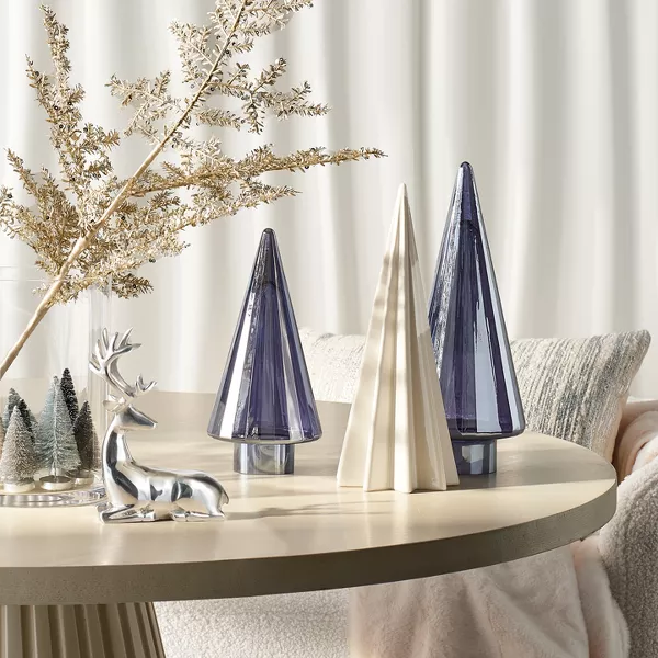 1 white ceramic & 2 metallic blue Christmas trees sit atop a table. A silver reindeer figurine sits in front of a large glass vase. Inside the vase, mini white & green Christmas trees line the bottom & large gold branches expand out. 