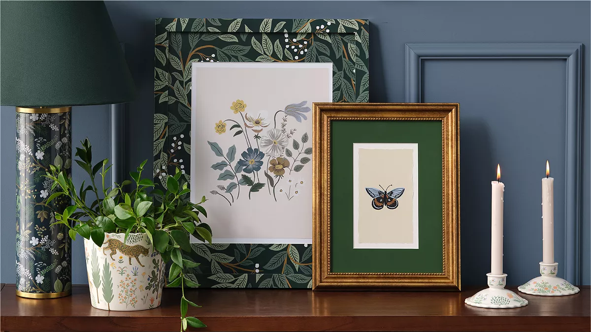 A shelf holds deep blue, black & green décor. There are 2 candlesticks with scalloped holders, a gold framed image of a butterfly, a larger floral framed image, a vine plant growing in a pot with forest imagery & a tall black lamp with floral designs.   