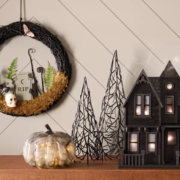 A black 2-story haunted house with lights sits on a table between a gold snake candle holder, 2 black metal trees & a clear glass pumpkin with twinkling lights inside. On the wall, a black twig wreath hangs showing a graveyard scene. 