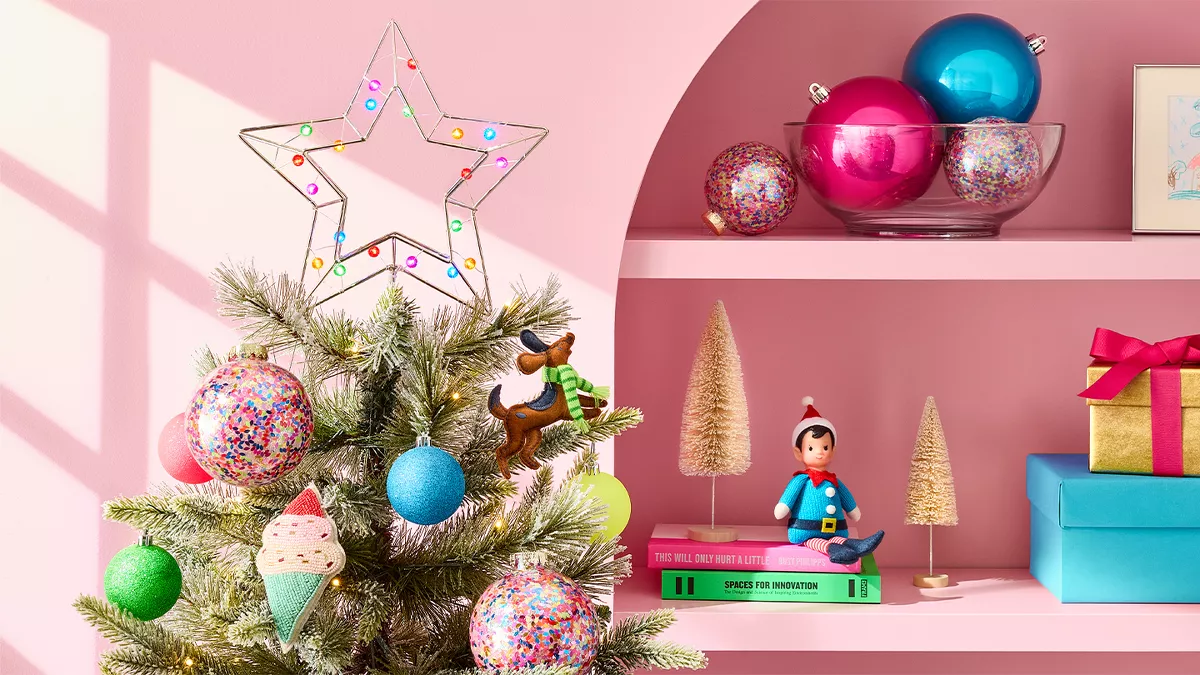 The image features a festive scene with a small Christmas tree decorated with colorful ornaments, such as round balls and an ice cream-shaped ornament. To the right, pink shelves hold more ornaments, small gold-colored trees, gift boxes, books, and a small toy figure. A child's drawing is also displayed. The walls are pink.