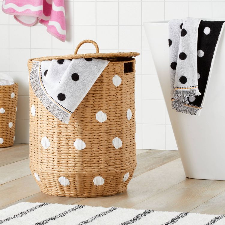 A woven basket with white polka dots and a lid, holding a fringed towel with black polka dots, placed on a wooden floor.