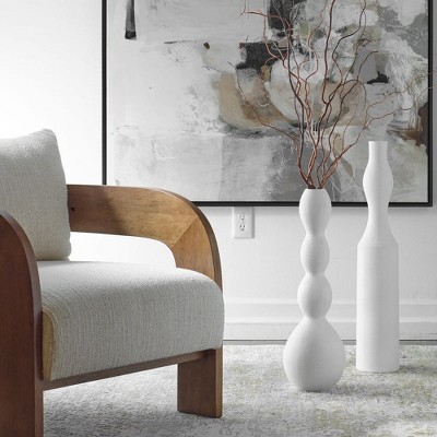 Two white colored vases with textured surfaces, one with a wavy design and branches inside, placed on a light-colored rug near a wooden armchair.