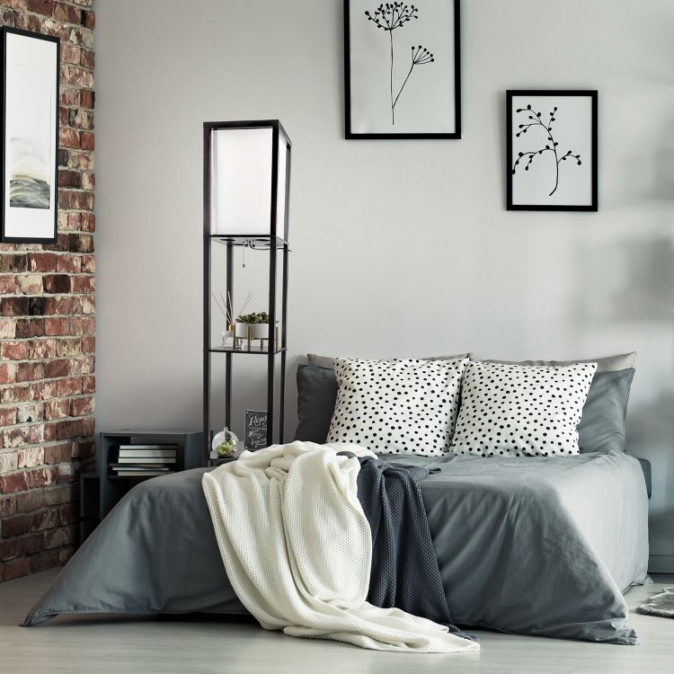 A tall black colored floor lamp with a white rectangular shade and shelves stands beside a bed with polka dot pillows.