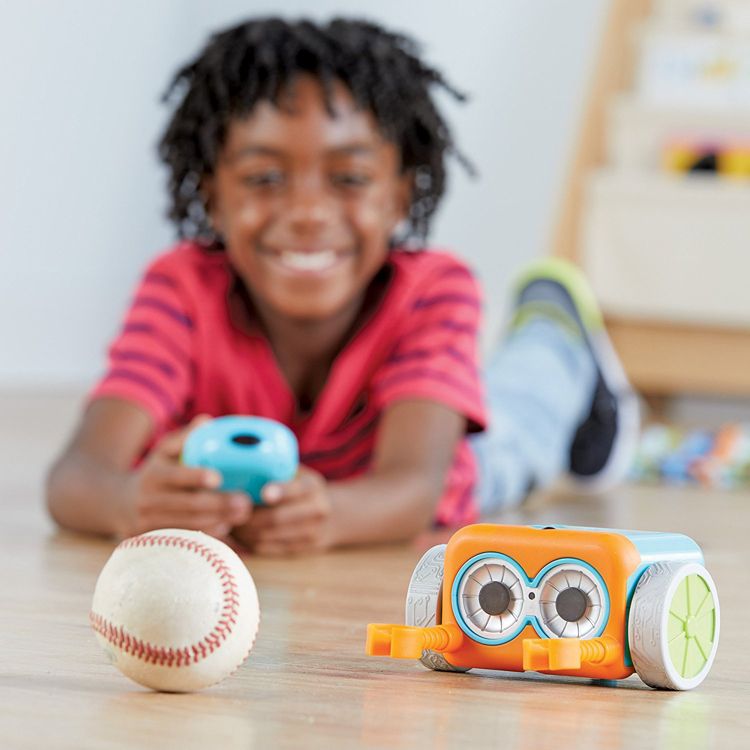 A small toy robot with an orange and blue body and large circular eyes is placed on a wooden floor near a baseball.