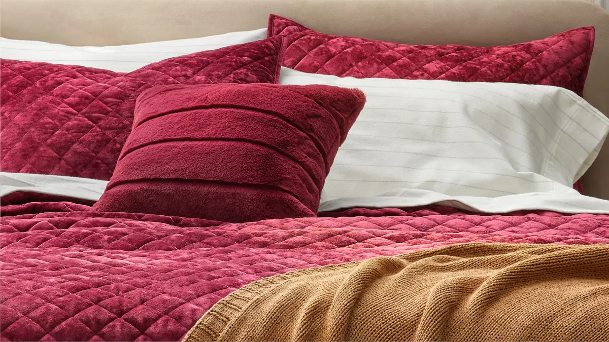 A sophisticated bedroom features a cream upholstered bed casually made with lush, cozy layers. Vibrant, berry-colored luxe velvet bedding is paired with flannel sheets & a velvet pillow to create a comfy & inviting bed.