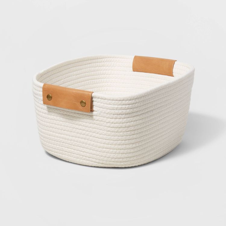 White coiled rope basket with a square base and tapered sides, featuring brown leather handles.