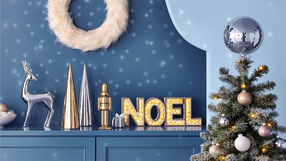 A festive scene with a blue backdrop. There is a fluffy white wreath, gold-colored cone decorations, a silver reindeer, and a sign with glowing "NOEL" letters. A small Christmas tree with baubles and a disco ball on top is on the right.