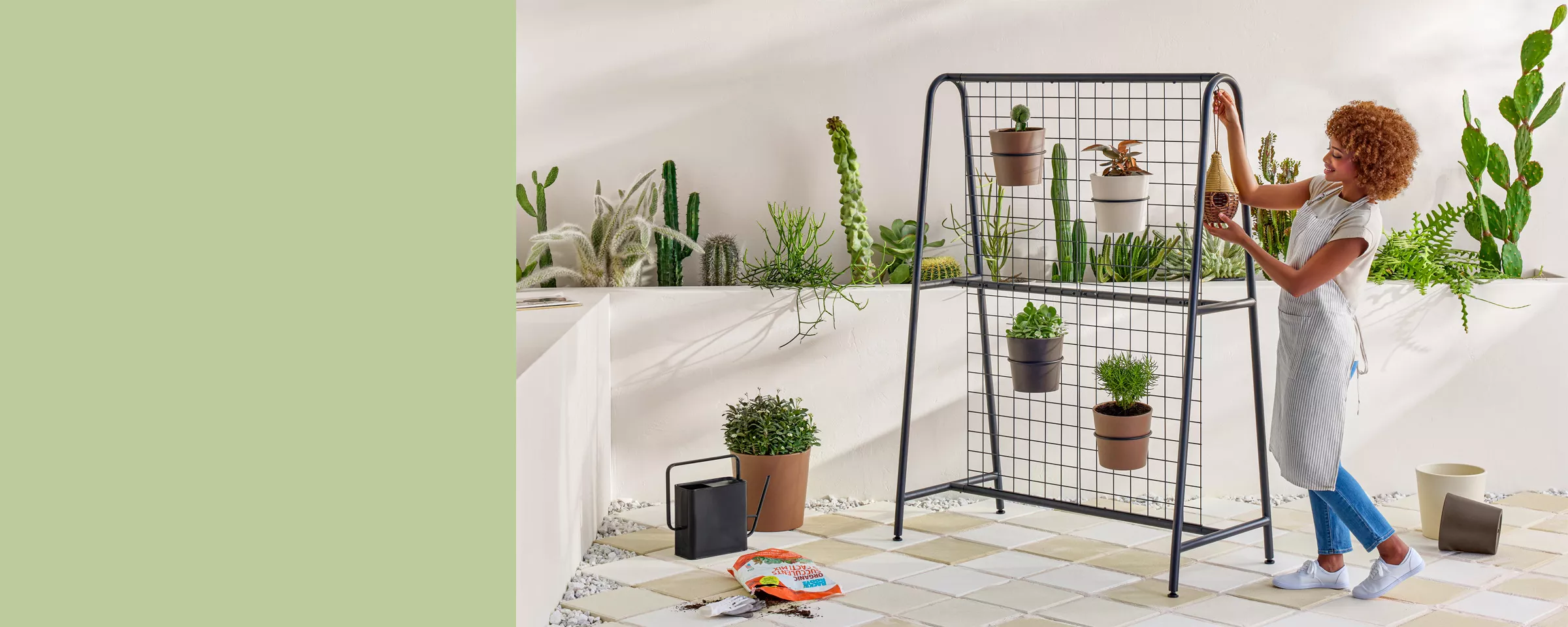 A lush outdoor patio is filled with various plants that pop against white walls & surroundings. In the middle, a decorative screen is adorned with more plants & a birdfeeder. An open bag of soil, watering can & pots are in the middle of being used. 